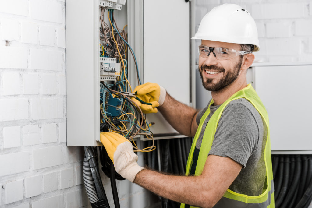 smiling electrician repairing electrical box with pliers in corridor and looking at camera